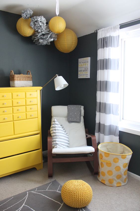 a black, white and yellow nursery with paper lamps and pompoms, a yellow dresser, a rocker, a basket and a knit ottoman plus striped curtains