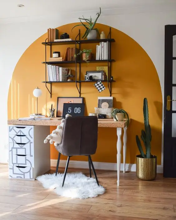 a boho working space with a color block yellow wall, a desk with a cork top, a cofy chair, an open shelving unit and some cacti