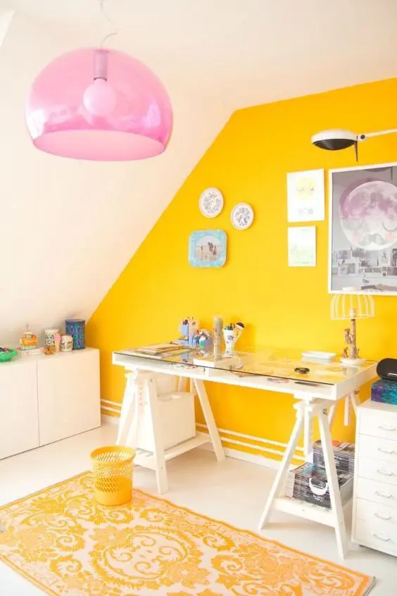a bold and shiny craft room with a sunny yellow accent wall, white furniture, a pink pendant lamp and pretty artworks