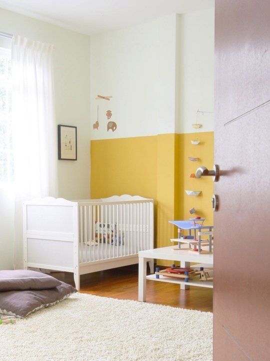 a bold nursery with a mustard and white color block wall, white furniture, colorufl toys and lilac pillows and white textiles