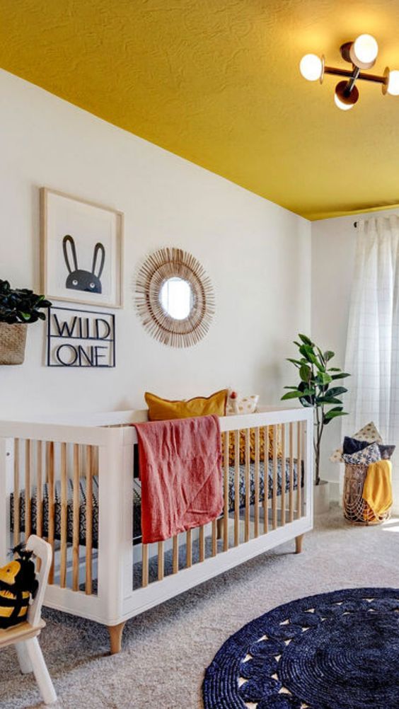 a bright and fun nursery with a yellow ceiling, some yellow linens, layered rugs, a crib and a pretty gallery wall in boho style