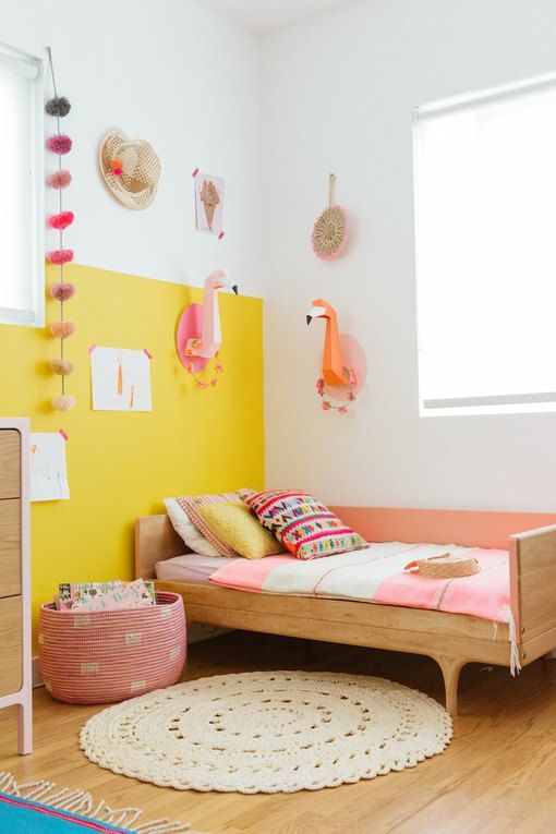 a colorful girl's room with a color block yellow wall, a simple bed and pink bedding, a pink basket, cardboard flamingo heads and other decor