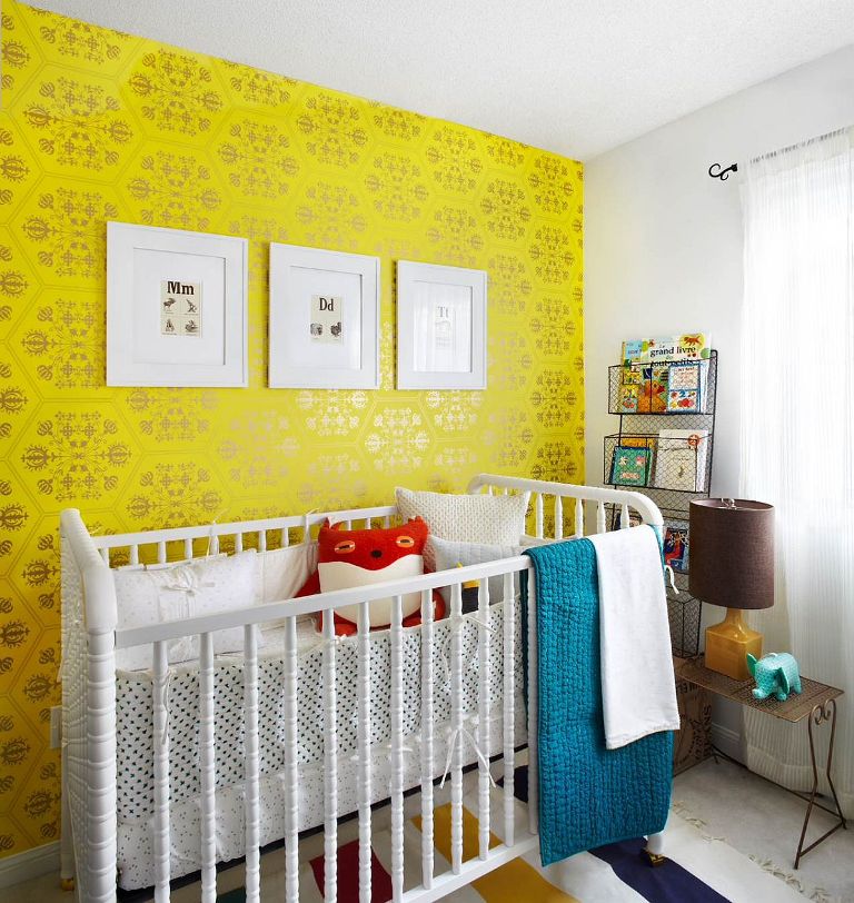 a colorful nursery with a bold yellow wallpaper wall, a white crib, wire shelf, colorful textiles and bedding plus a gallery wall