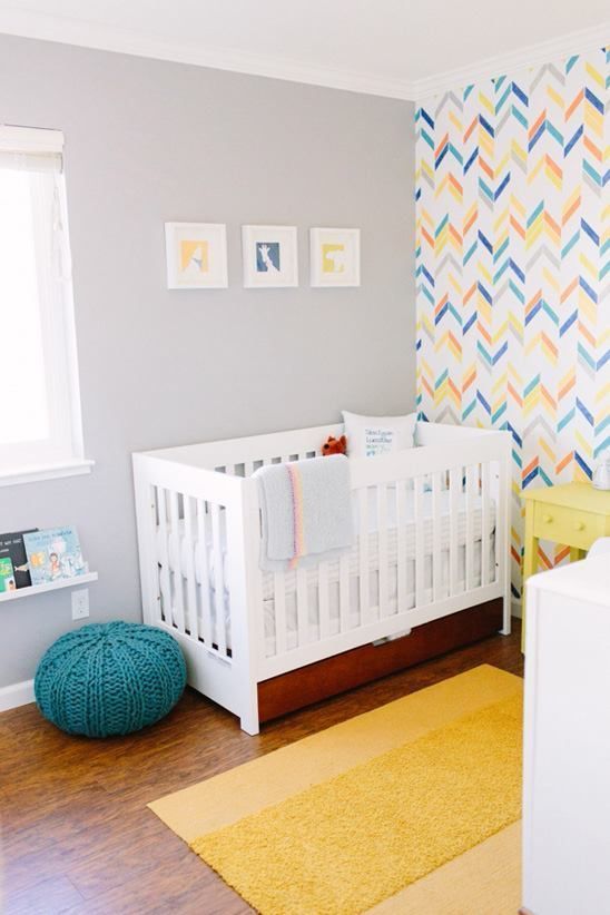 a colorful nursery with a bright geometric wall, white and yellow furniture, a yellow rug, a blue ottoman and a gallery wall