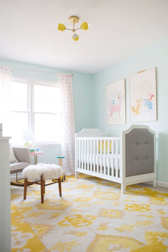 a colorful nursery with light blue walls, neutral furniture, a colorful gallery wall, a printed rug and curtains plus a yellow chandelier