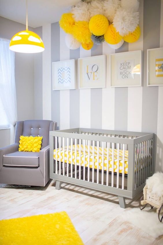 a pretty grey, white and yellow nursery with a striped accent wall, grey furniture, lots of paper pompoms and a yellow pendant lamp
