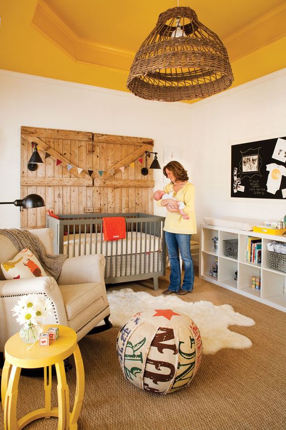 a rustic nursery with a yellow ceiling, grey and white furniture, a wooden door, a colorful ottoman and yellow touches