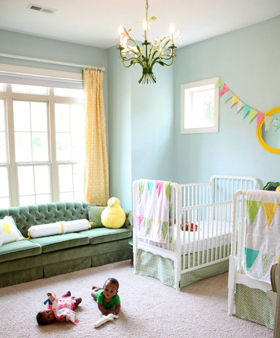 a shared nursery with light blue walls, a green sofa, white cribs, a colorful banner and bedding, bold yellow touches and a floral chandelier