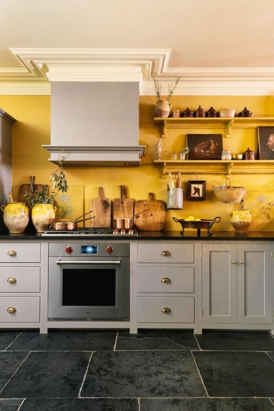 a vintage kitchen with grey cabinetry, black countertops, a yellow accent wall and open shelves with pretty artworks