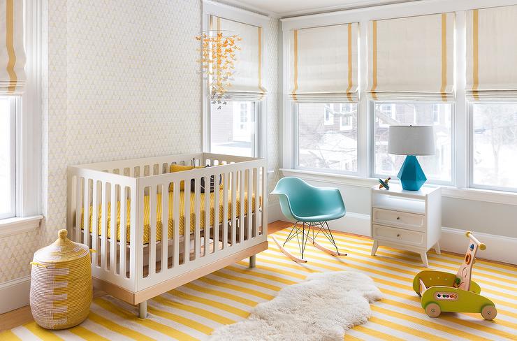 an airy nursery with white walls, a striped yellow rug, a crib with yellow bedding, some toys and Roman shades with yellow stripes