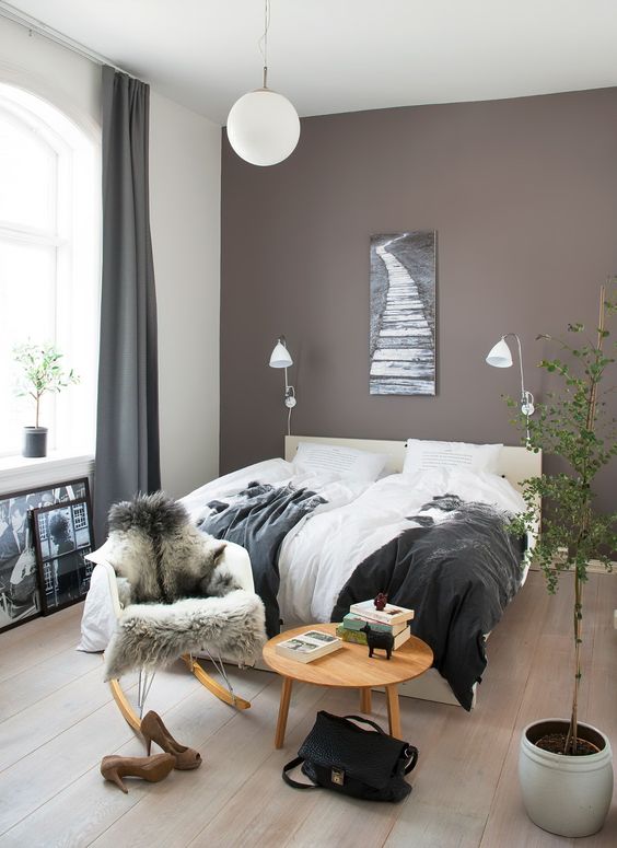a Scandinavian bedroom with a mauve accent wall, a creamy bed, a chair and a table, sconces and some potted greenery