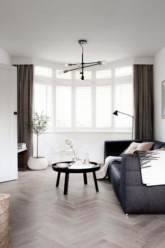 a Scandinavian living room with a bow window, a black sofa, a round table and taupe curtains paired with shutters