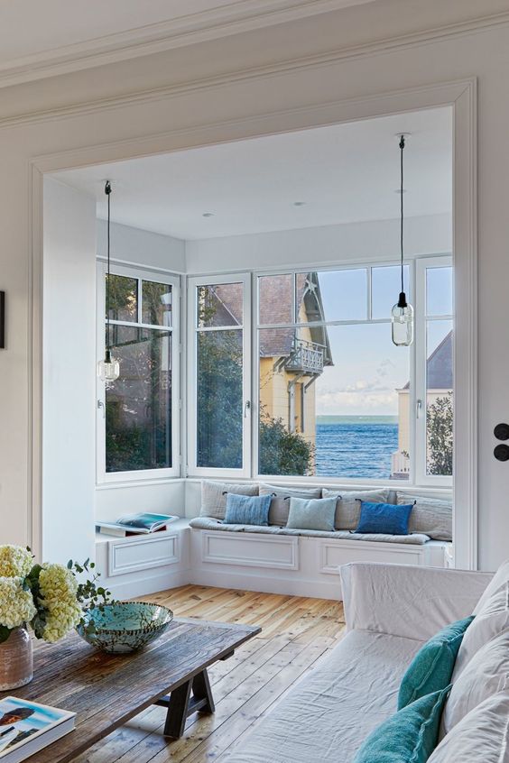 a beautiful coastal living room with a bow window that shows off amazing sea views and a windowsill bench with pillows
