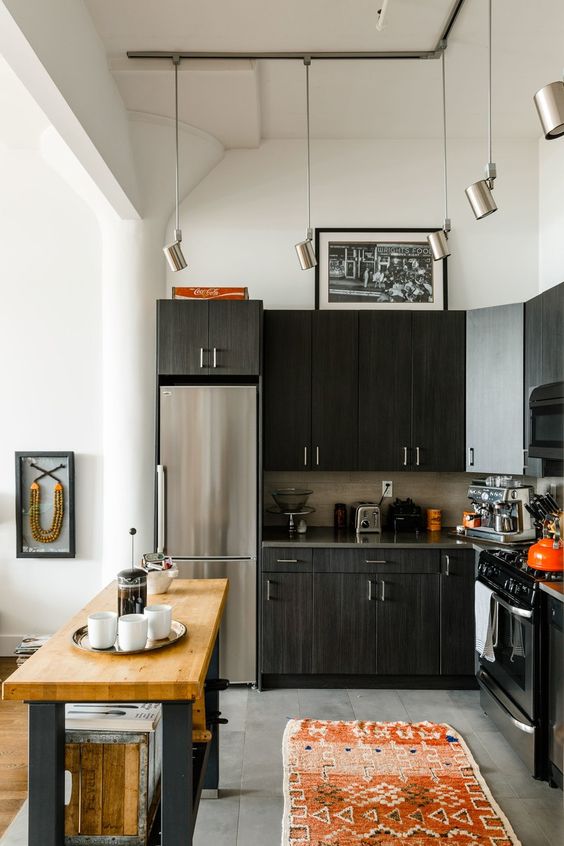 A black L shaped kitchen with a tile backsplash, a small kitchen island with a butcherblock countertop and pendant lamps