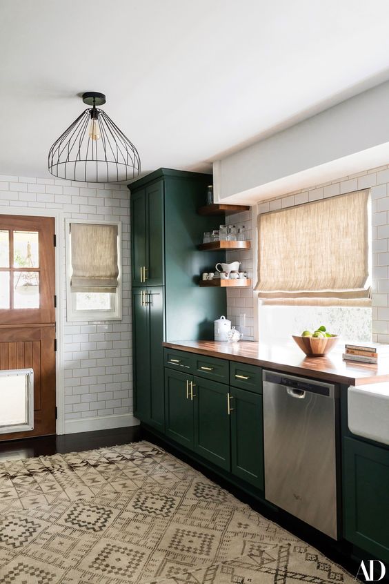 a chic dark green farmhouse kitchen with a buctcherblock countertop, woven shades and metal pendant lamps plus a boho rug