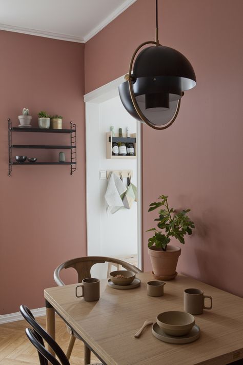 a chic dining room with mauve walls, a wooden table and mismatching chairs, a pendant lamp and an open shelving unit