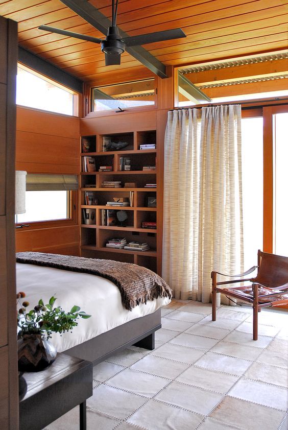 A cozy mid century modern bedroom with a built in shelving unit, a bed and a leather chair and some clerestory windows