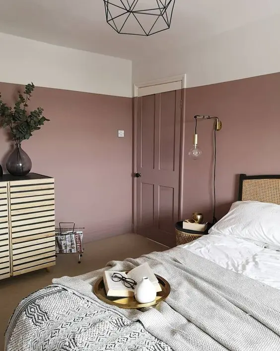 a lovely bedroom with mauve walls, a rattan bed, a wooden sideboard, neutral bedding and touches of gold