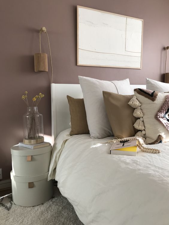 a mauve bedroom with a white bed with neutral boho bedding, stacked boxes, wooden sconces and a statement artwork