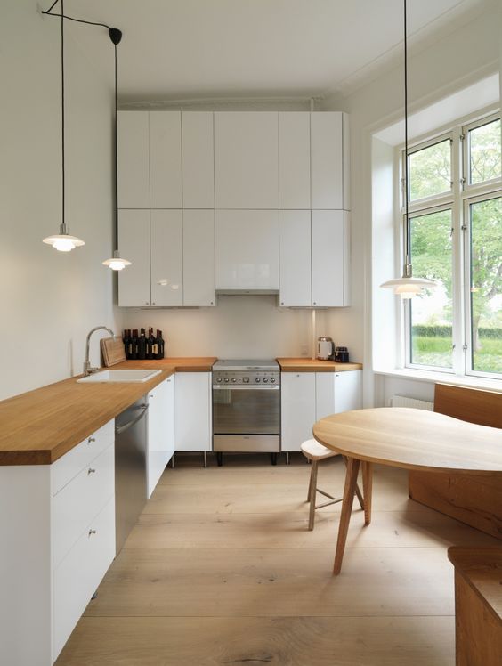 a modern white L-shaped kitchen with butcherblock countertops and a sleek white backsplash plus a dining space by the window