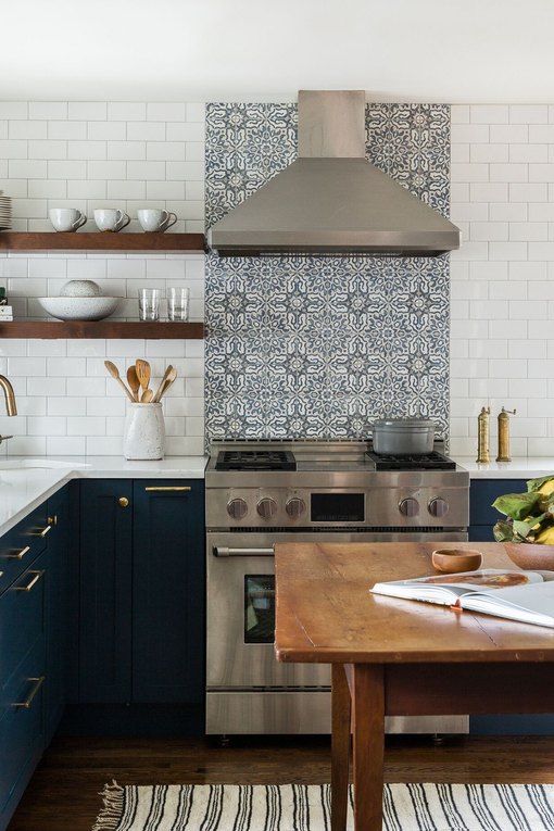 A navy L shaped kitchen with a white and patterned tile backsplash and a wooden table as a kitchen island or for having meals