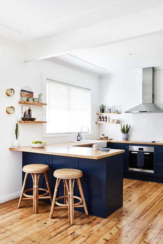 A navy U shaped kitchen with light stained butcherblock countertops, a white tile backsplash and rattan stools