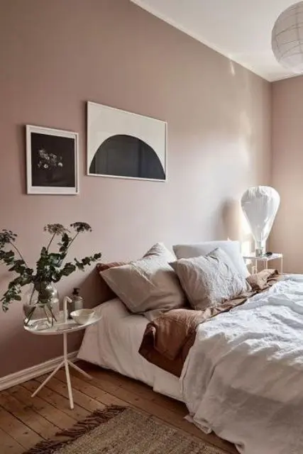 a pretty Scandinavian bedroom with mauve walls, a comfy bed and neutral bbedding, mismatching nightstands and cool artworks
