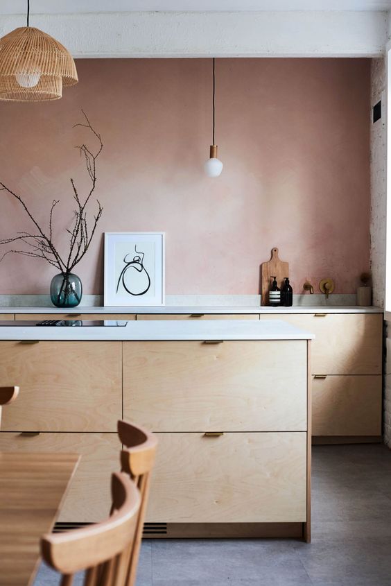 a pretty kitchen with a mauve accent wall, plywood cabinets, white countertops and pendant lamps