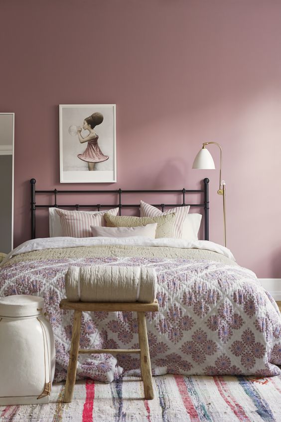 a pretty mauve bedroom with a black forged bed, neutral and pastel bedding, a stool, a sconce and a cool artwork