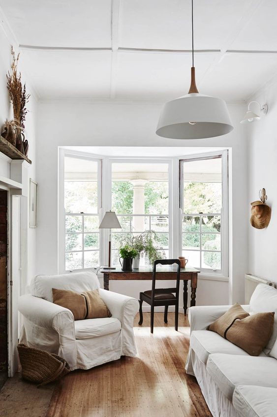 A refined neutral living room with white furniture, rust colored pillows,a  vintage desk and chair by the bow window