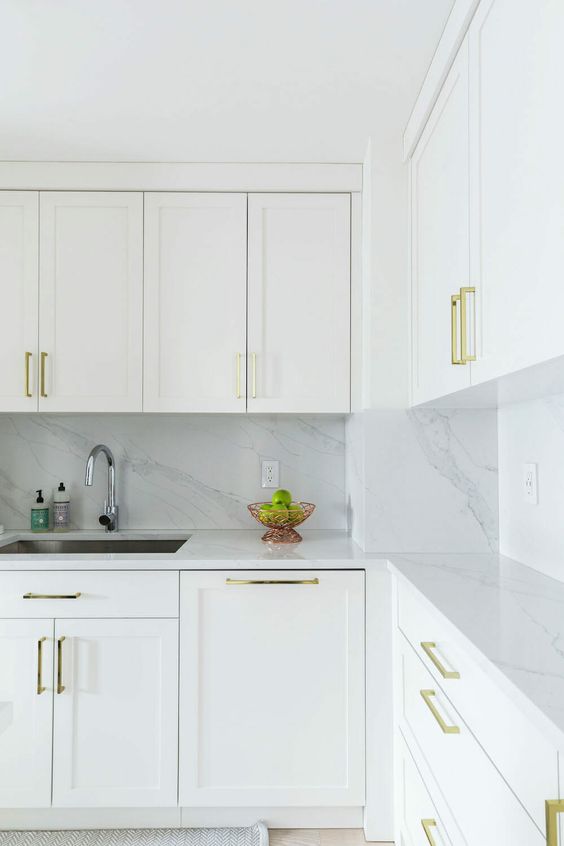 a serene white kitchen with shaker cabinets, a white marble backsplash and countertops plus gold handles is a chic space