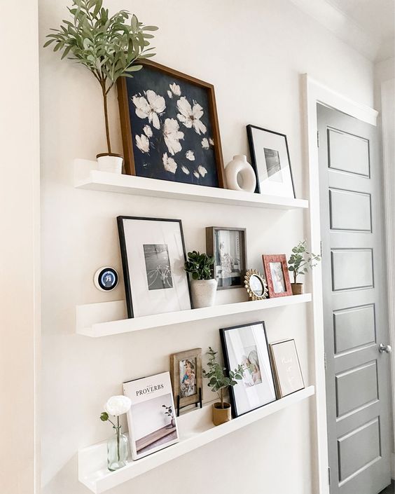 a small farmhouse gallery wall with white ledges, pretty artworks, potted greenery, vases and a photo is a chic idea