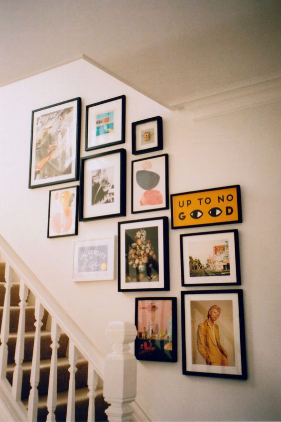 a small gallery wall with mismatching black frames and pretty and bright artworks and other stuff is a cool idea to take the space over the stairs