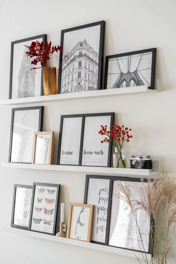a stylish gallery wall with white ledges, art in black frames, dried blooms in vases, a candle and a vintage camera is a cool idea for a Scandi space