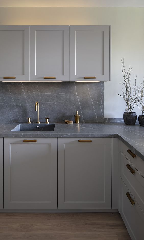 a stylish monochromatic grey kitchen with shaker style cabinets, a grey marble backsplash and countertops plus brass fixtures and handles