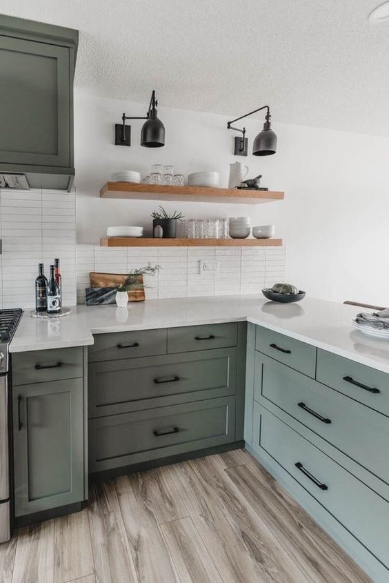 a stylish sage green kitchen with shaker style cabinets, white countertops and a white skinny tile backsplash plus floating shelves