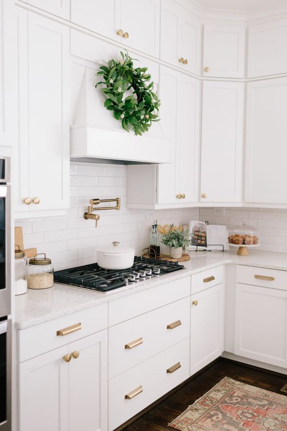 a white farmhouse kitchen with shaker style cabinets, white quartz countertops and a white skinny tile backsplash plus brass fixtures