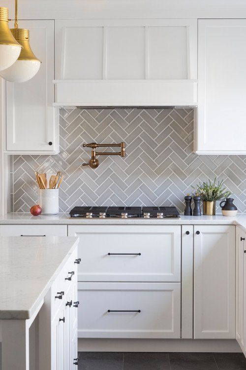 an exquisite white kitchen with shaker cabinets, white quartz countertops, a grey herringbone tile backsplash and touches of gold and brass
