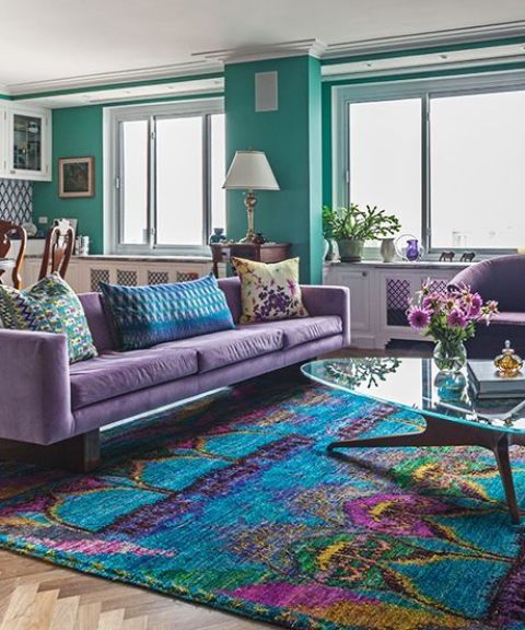 a bright living room with green walls, a purple sofa and chairs, a bright rug with purple touches and a glass coffee table