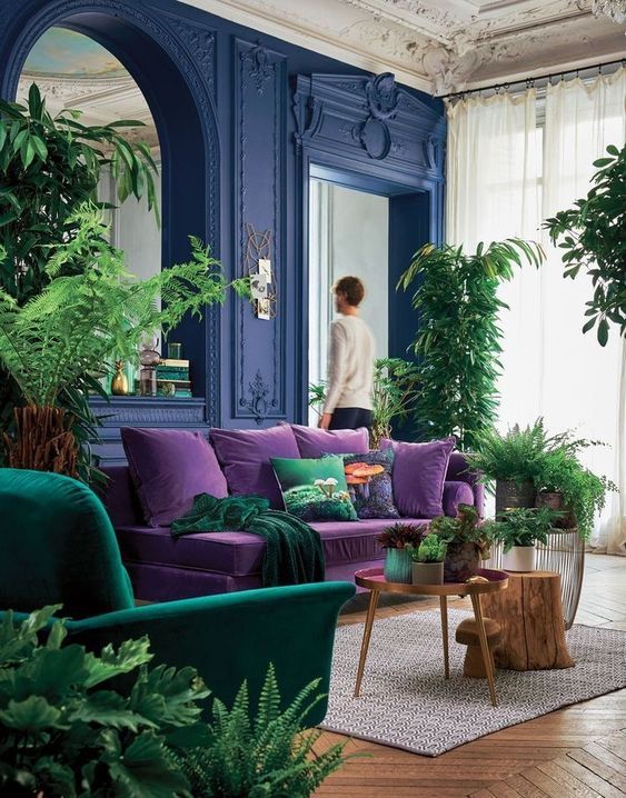 03 a bright living room with bold blue walls, a purple sofa, an emerald chair, potted greenery and a large mirror