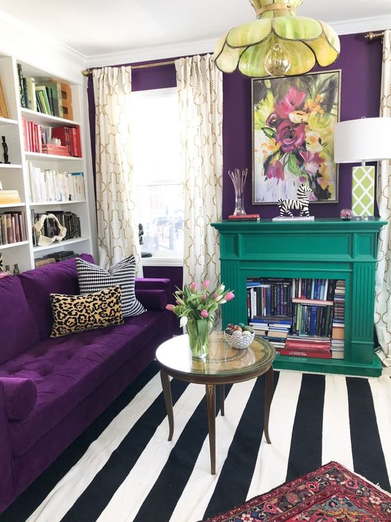 a colorful living room with a depe purple accent wall and a matching sofa, a striped rug, a floral pendant lamp and an emerald fireplace with books