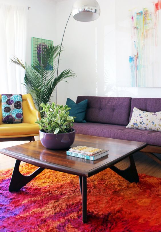 a colorful living room with a purple sofa, a yellow chair, a chic stained coffee table and bright artworks and a floor lamp