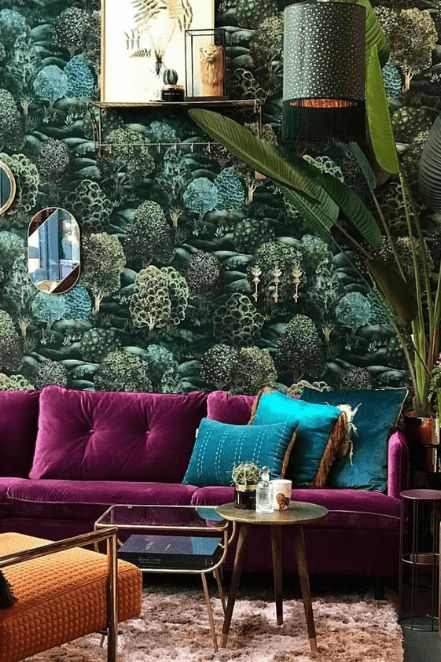 a maximalist living room with a botanical accent wall, a purple sofa, green pillows, a pendant lamp and a potted plant plus mismatching tables