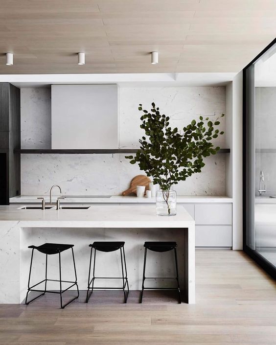 a minimalist off-white kitchen with sleek cabinets, a hood that matches them and black stools and a white stone backsplash