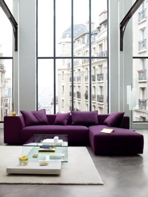 a contemporary living room done in neutrals, with double height windows, a purple sectional, a glass table and a neutral rug