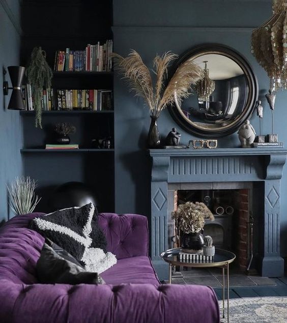 a moody living room with slate grey walls, a fireplace, a purple tufted sofa, a round mirror and a mother of pearl chandelier
