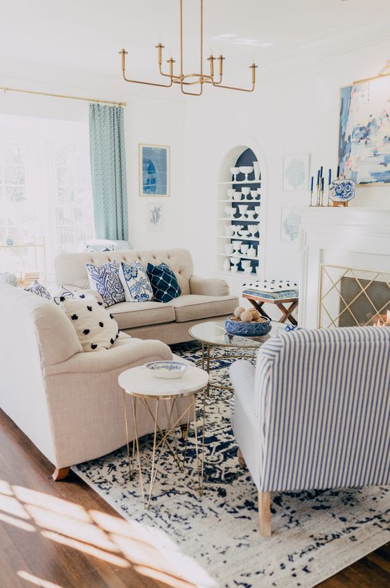 a catchy living room with a fireplace, neutral sofas, blue printed pillows and a striped chair, an elegant chandelier and a blue niche with tableware