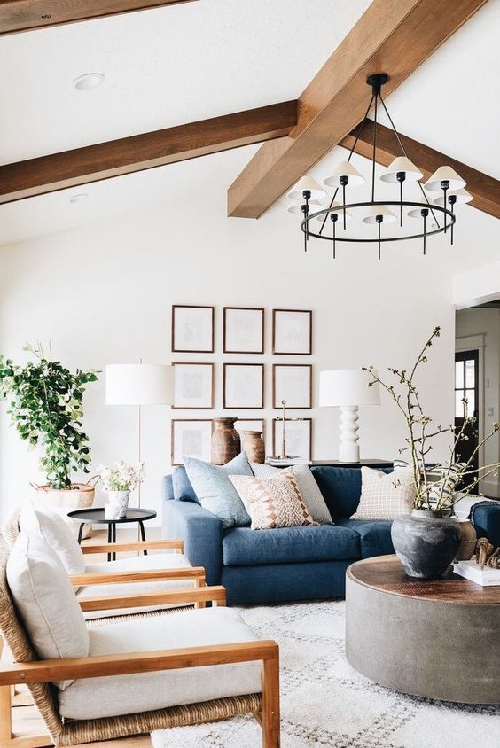 a chic farmhouse living room with wooden beams, a navy sofa, neutral chairs, a round coffee table and a gallery wall
