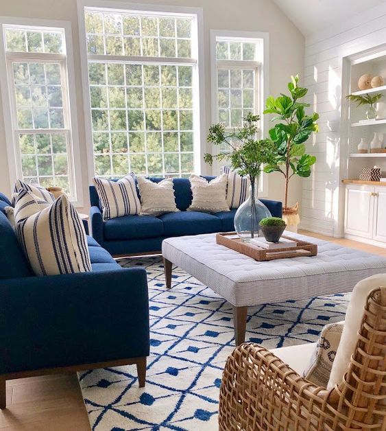 A beautiful coastal living room with navy sofas, a striped ottoman, white built in storage units and a white rattan chair