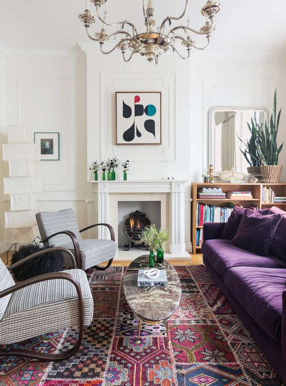 An eclectic living room with a non working fireplace, a depe purple sofa, printed chairs, a refined chandelier and a bold printed rug